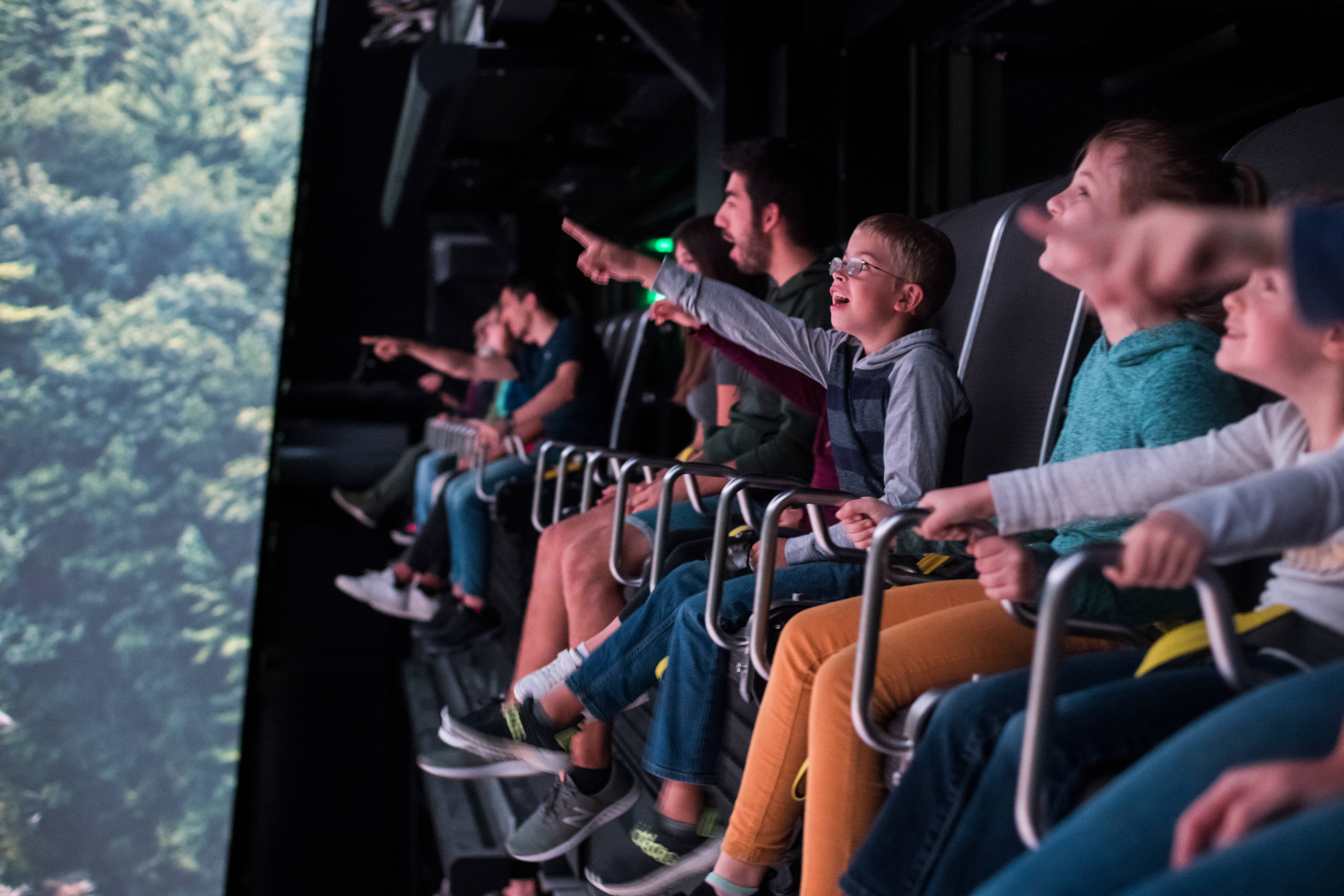 Take Flight Adventure Fly Ride Open to Public. Website featured image for sharing shows a group of people secured in seats riding Take Flight the attraction while their feet dangle over the edge.